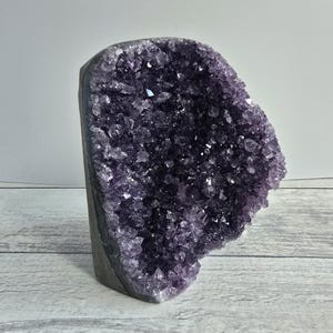Può includere: Una grande geode di ametista grezza, di colore viola intenso. La formazione cristallina è visibile, con numerosi piccoli punti scintillanti. La geode è posta su una superficie chiara, mettendo in risalto la sua bellezza naturale. Un oggetto decorativo.