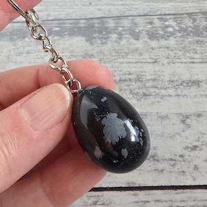 May include: A black snowflake obsidian gemstone keyring. The polished, oval-shaped stone has white and grey snowflake-like patterns. The keyring has a silver-coloured chain and clasp.