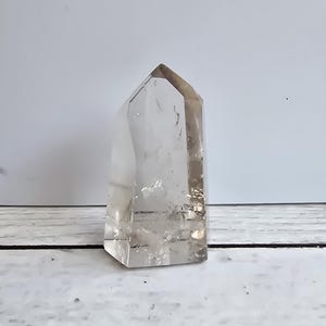 Op de afbeelding: Een heldere kwartskristalpunt met een facetgeslepen bovenkant. Het kristal is transparant met subtiele interne insluitsels en staat op een wit en grijs houten oppervlak. Het kristal is een natuurlijk, decoratief item.
