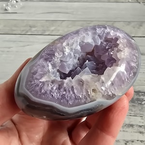 Pode incluir: Uma geodo de ametista polida, mostrando uma cavidade preenchida com cristais roxos brilhantes. A casca externa da geodo é uma mistura de faixas cinza e brancas, criando um contraste marcante com o interior vibrante. A geodo é segurada em uma mão.