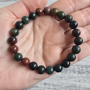 Puede incluir: Una pulsera de cuentas con piedras preciosas redondas y pulidas en tonos verde oscuro, marrón y burdeos. Las cuentas están ensartadas en una banda elástica. La pulsera se muestra sobre una superficie clara.