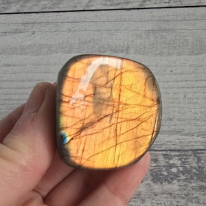 Può includere: Una pietra preziosa di labradorite lucidata, di forma rettangolare, con una calda tonalità arancione dorato. La pietra presenta intricati motivi scuri, simili a venature, e un sottile lampo blu. La pietra è tenuta in mano.
