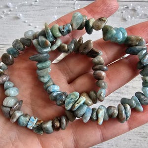 May include: Two beaded bracelets made of irregular, polished stones in shades of blue, green, and brown. The bracelets are held in a hand, showcasing the natural variations in the stones. The bracelets are a jewelry item.