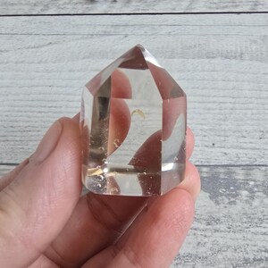 May include: A clear quartz crystal point with a faceted design. The crystal is held in a hand, highlighting its transparency and internal features. The crystal's shape is a truncated pyramid, suitable for meditation and energy work.