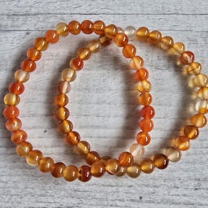 Può includere: Due braccialetti di perline arancioni e gialle. Le perline sono rotonde e hanno un aspetto lucido e traslucido. I braccialetti sono fatti di piccole perline uniformi, infilate insieme per formare una fascia flessibile. La gradazione di colore va dal giallo chiaro all'arancione intenso.