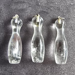 Könnte beinhalten: Drei klare, kegelförmige Anhänger aus transparentem Material. Jeder Anhänger hat ein abgerundetes Oberteil, einen schmalen Hals und eine breitere Basis, mit einem kleinen Metallring an der Oberseite zum Aufhängen. Das Innere jedes Stücks weist sichtbare Einschlüsse auf.