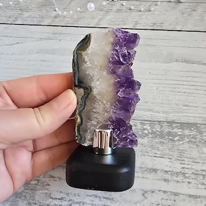 Peut inclure: Un ornement en cristal d'améthyste avec un dessus argenté et une base noire. Le cristal présente un dégradé de couleurs, dont le blanc, le gris et le violet profond. La base est de forme carrée.