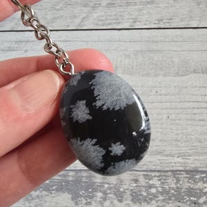 Op de afbeelding: Een zwarte en witte sneeuwvlok obsidiaan stenen sleutelhanger. De ovale steen heeft een gepolijst oppervlak met grijze sneeuwvlokachtige patronen. De sleutelhanger heeft een zilverkleurige ketting en ring.
