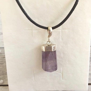 Könnte beinhalten: Ein lila Amethyst-Anhänger mit silberner Kappe hängt an einer schwarzen Kordel. Der Edelstein hat eine rechteckige Form mit facettierter Unterseite. Der Anhänger wird vor einem weißen Hintergrund präsentiert.