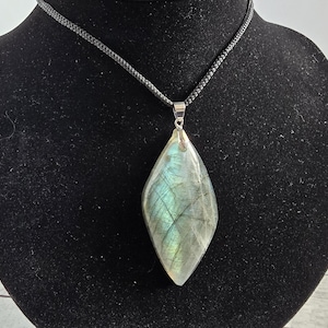 Pode incluir: Colar com pingente em forma de gota com uma pedra de labradorita brilhante e iridescente. A pedra exibe tons de azul, verde e cinza. O pingente é suspenso por um colar de cordão preto com fecho prateado.