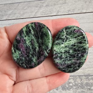 Può includere: Due pietre ovali di zoisite con finitura lucida. Le pietre mostrano un mix di tonalità verdi, nere e viola. Le pietre di zoisite sono tenute in una mano, mostrando i loro colori e le loro trame naturali.