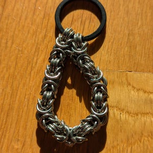 May include: A handcrafted keychain with a silver-coloured chainmail design in a teardrop shape, attached to a black rubber ring. The chainmail is intricately woven, creating a textured appearance. The keychain is displayed on a wooden surface.