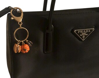 May include: Black handbag with a gold-tone keychain featuring miniature pumpkin, macaron, and coffee bean charms. The bag has a triangular Prada logo and a top handle.
