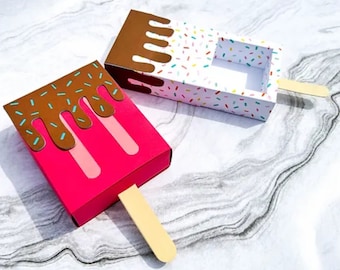 May include: Two popsicle shaped boxes with a stick handle. One box is pink with brown chocolate dripping down the side and sprinkles. The other box is white with brown chocolate dripping down the side and colorful sprinkles.