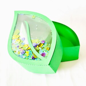 May include: A green leaf-shaped box with a clear plastic window. The window is filled with colorful sequins and glitter. The box is open and the lid is lying next to it.