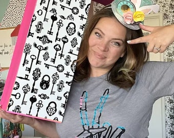 May include: A woman wearing a gray t-shirt with a skeleton hand peace sign design and a whimsical Alice in Wonderland themed headband. The headband features a clock, a bunny ear, and a flower.