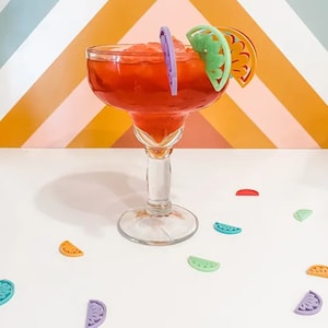 May include: A red margarita cocktail in a glass with a lime wedge garnish. The drink is surrounded by colorful plastic fruit slices.