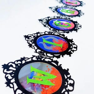 May include: A series of decorative oval-shaped embellishments with black filigree frames. Each oval features a letter in bright green against a colorful background of blue, red, and silver. The letters appear to spell a word.