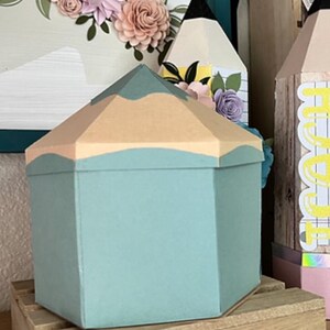 May include: A teal blue pencil-shaped box with a light brown top. The box is sitting on a wooden base with the words "@craftyhobbysbybee" printed on it.