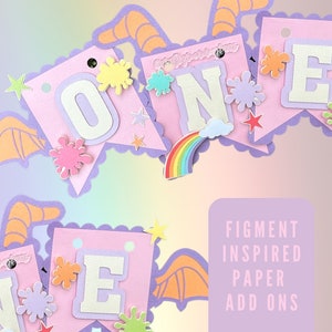 Pode incluir: Uma faixa de papel rosa com a palavra "ONE" em letras brancas, decorada com respingos de tinta coloridos, estrelas e um arco-íris. A faixa tem uma borda ondulada e faz parte de um conjunto "Figment Inspired Paper Add Ons".