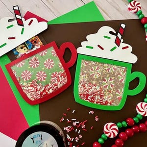 Könnte beinhalten: Zwei grüne und rote Weihnachts-Heißgetränkebecher mit weißer Schlagsahne und Streuseln. Die Becher sind aus Papier und haben ein transparentes Plastikfenster, um die Streusel im Inneren zu zeigen.