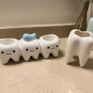 May include: A set of white ceramic tooth-shaped containers. One is a single tooth, and the other is a set of three teeth with smiling faces. The middle tooth has a blue toothpaste accent. These are likely toothbrush holders or decorative items.