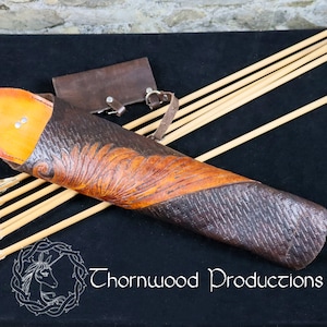 May include: A brown leather arrow quiver with a tooled design of a stylized bird wing. The quiver is on a black surface with several arrows with fletching.