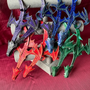 May include: Five leather dragon head keychains in different colours: purple, blue, red, green, and green. The keychains are displayed on a grey stand.