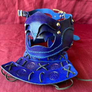 May include: A blue leather mask with a pointed snout and decorative stitching. The mask is attached to a blue leather collar with a buckle. The mask is accompanied by three blue leather straps with Celtic knot designs and black laces.