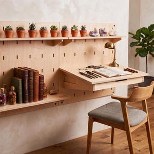 Könnte beinhalten: Ein helles, an der Wand montiertes Lochplattensystem aus Holz mit einem modularen Schreibtischaufsatz, mehreren horizontalen Regalen und einem passenden Holzstuhl mit grauem Stoffbezug. Die Regale enthalten kleine Sukkulenten, Bücher und Glasgefäße.