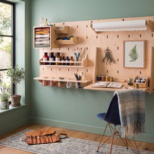 Op de afbeelding: Een wandorganizer van lichtgekleurd hout met een geperforeerde plaat, een uitklapbaar bureau, een papierrolhouder, diverse planken en opbergvakken. De unit bevat diverse kunstbenodigdheden, glazen potten, een ingelijste varenprint en gereedschap.