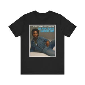 Op de afbeelding: Zwart T-shirt met een rechthoekige afbeelding. De afbeelding toont de tekst "AFRICAN AMERICAN EAGLE OUR PEOPLE ALWAYS HAD GREAT JEANS" en een man in een spijkerjas en jeans.