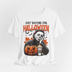 Könnte beinhalten: Weißes T-Shirt mit einem Motiv eines Charakters aus einem Horrorfilm, der einen Kürbis und ein Kaffeegetränk hält. Der Text "Just Waiting for Halloween" ist auf dem Shirt aufgedruckt.
