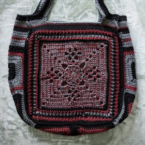 May include: A crocheted tote bag with a large, intricate, red, black, and gray square design in the center. The bag has a wide, black strap.