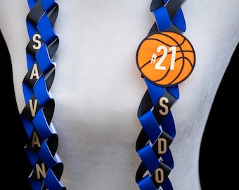 Puede incluir: Un collar de flores azul y negro con el nombre "SAVANNAH" y "SDHS" escrito en letras doradas. Una pelota de baloncesto con el número "21" está unida al collar.