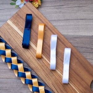 May include: A wooden cutting board displays a selection of ribbons in navy blue, gold, and white. A woven ribbon in navy and gold is also present. Decorative paper flowers in red, yellow, and blue are in the background.