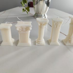 May include: Five white pillar candles with intricate details. The candles are arranged in a row on a white surface.