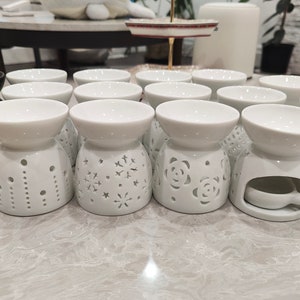 May include: A set of 10 white ceramic oil diffusers with various decorative cutouts. The diffusers have a round top and a wide base with a small opening in the centre.