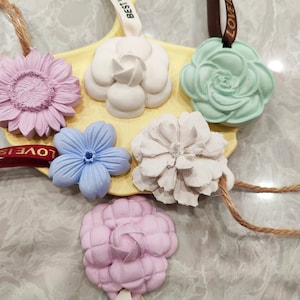 May include: Six different flower-shaped air fresheners with strings for hanging. The flowers are in shades of purple, blue, green, and white. The flowers are on a yellow surface with the words "LOVE IS BETTER" and "LOVE" printed on the strings.