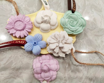 May include: Six different flower-shaped air fresheners with strings for hanging. The flowers are in shades of purple, blue, green, and white. The flowers are on a yellow surface with the words "LOVE IS BETTER" and "LOVE" printed on the strings.