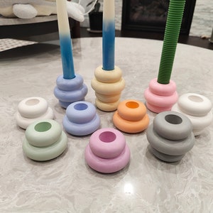 May include: A set of nine stacked candle holders in various pastel colors, including light blue, pink, green, gray, and white. The holders are arranged in a circle on a light gray surface.