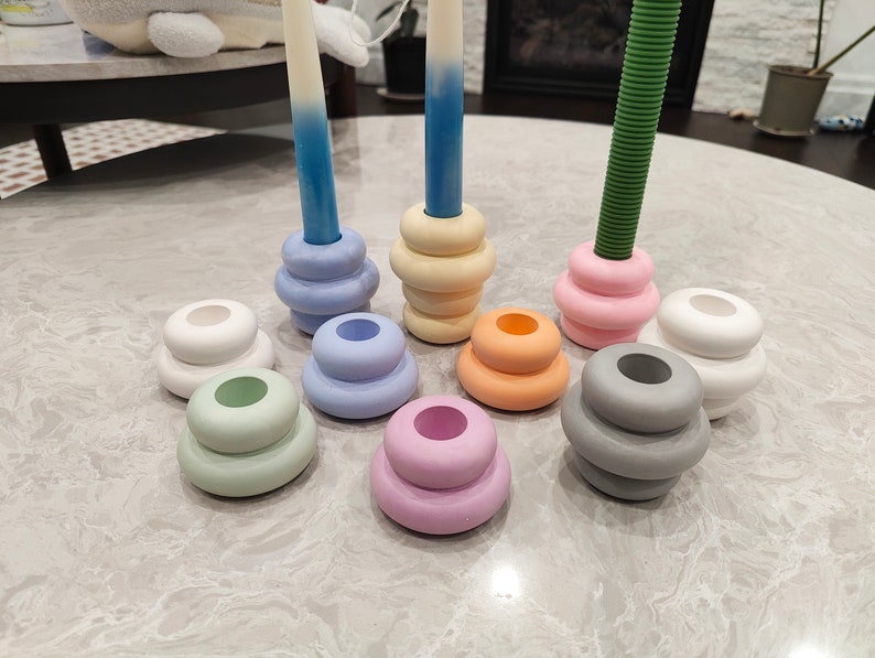 May include: A set of nine stacked candle holders in various pastel colors, including light blue, pink, green, gray, and white. The holders are arranged in a circle on a light gray surface.