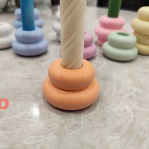 May include: A set of five candle holders in various colors, including white, light blue, pink, green, and yellow. Each holder is made of ceramic and has a stacked design. The candle holders are arranged on a white marble surface.