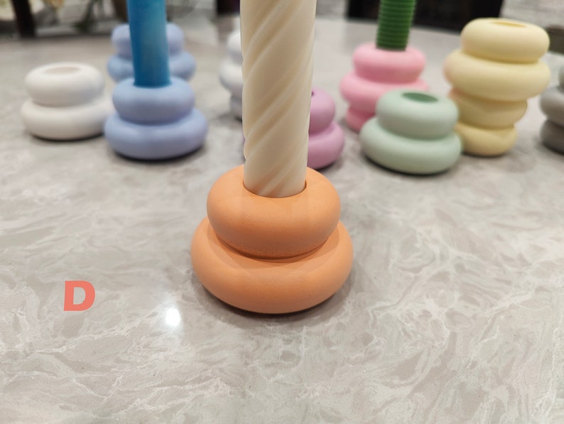 May include: A set of five candle holders in various colors, including white, light blue, pink, green, and yellow. Each holder is made of ceramic and has a stacked design. The candle holders are arranged on a white marble surface.