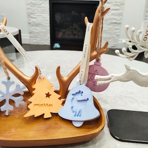 May include: A wooden tray with a gold metal stand holds five Christmas ornaments. The ornaments are a snowflake, a Christmas tree, a bell, a reindeer, and a round ornament with a snowflake design. The ornaments are decorated with the words "I miss you" and "merry".