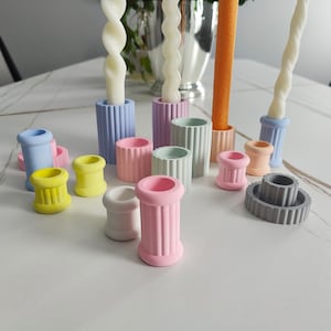 May include: A set of colorful candle holders made of concrete. The holders are in various pastel colors, including light blue, pink, yellow, and gray. The holders are arranged on a white table.