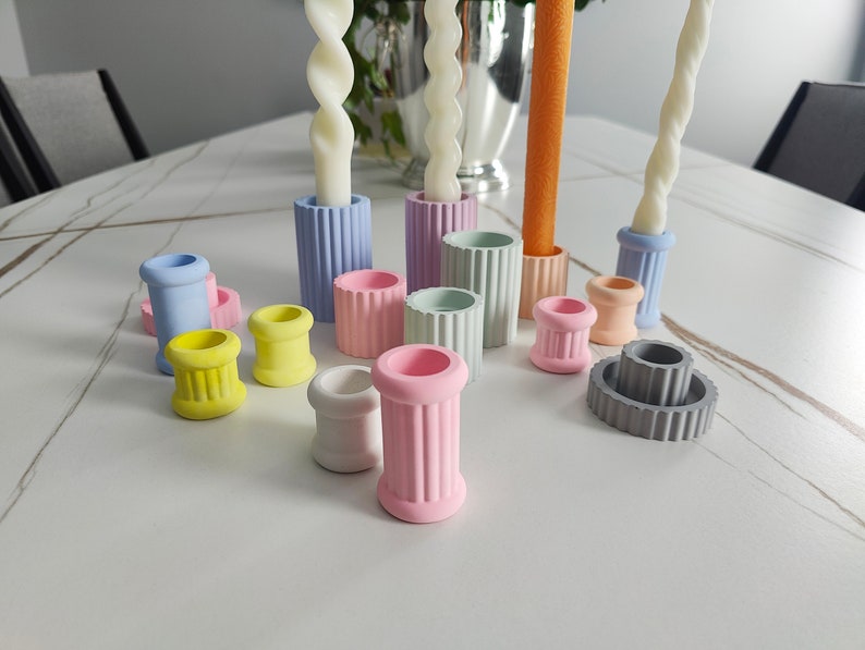 May include: A set of colorful candle holders made of concrete. The holders are in various pastel colors, including light blue, pink, yellow, and gray. The holders are arranged on a white table.