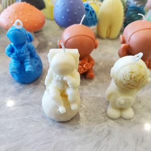 May include: A collection of astronaut-themed candles, including a blue astronaut sitting on the moon, a white astronaut holding a flower, and a white astronaut with gold glitter on its helmet.