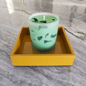 May include: A green candle in a frosted glass container sits on a mustard yellow square tray. The candle has a white wick and is partially melted.