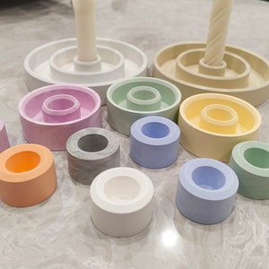 Peut inclure: Un ensemble de 10 bougeoirs ronds de différentes couleurs pastel, dont rose clair, bleu clair, vert clair, gris et blanc. Les bougeoirs sont disposés en cercle sur une surface gris clair.